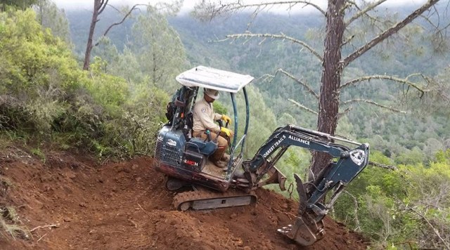 A trail crew member operating a mini excavator to cut new tread across a steep red-dirt hillside
