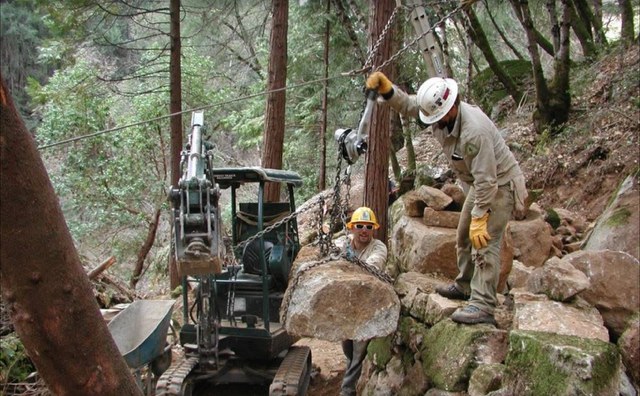 Crew members guiding a large boulder hung from an excavator into a stacked rock retaining wall
